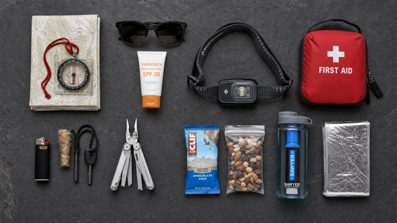 Flat-lay of the ten essentials for hiking: map and compass, sunscreen, headlamp, first-aid kit, fire starter, multi-tool, snacks, water filter and emergency blanket